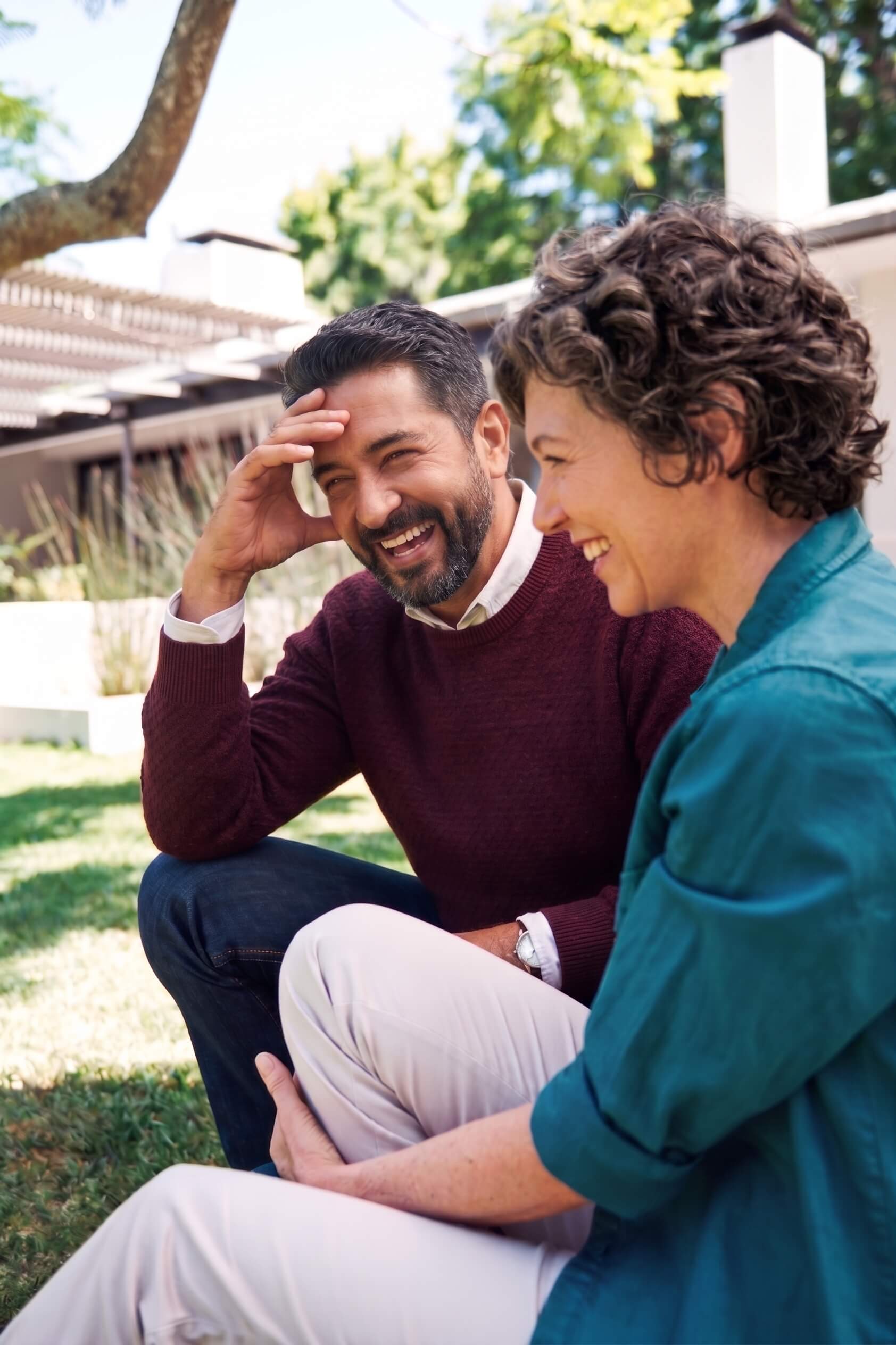 A couple sits outdoors on bright green grass. They laugh together. The man wears a maroon sweater and rests his hand on his head. The woman smiles beside him. She has short gray curls and wears a teal shirt. Their posture is relaxed. This joyful connection reflects a regulated nervous system.